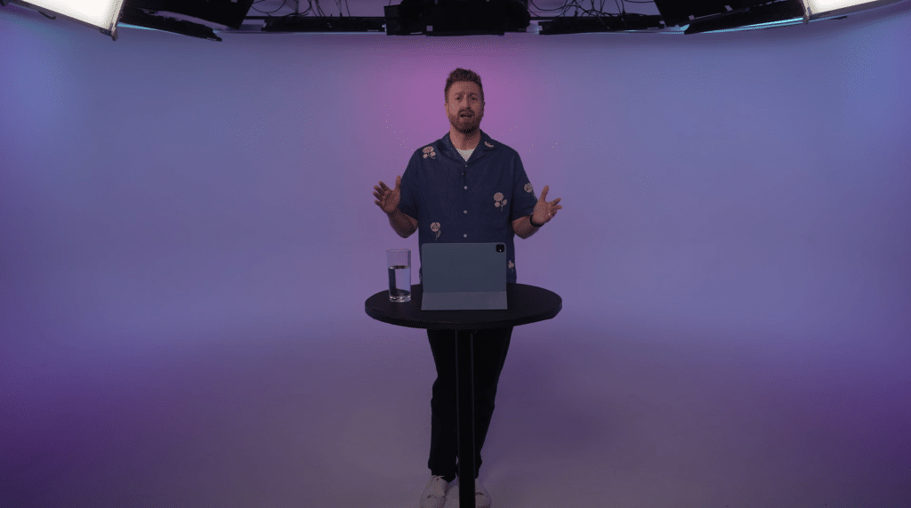 A man stands behind a round black table with a laptop and a glass of water, speaking in a studio with a purple gradient background and overhead lighting.